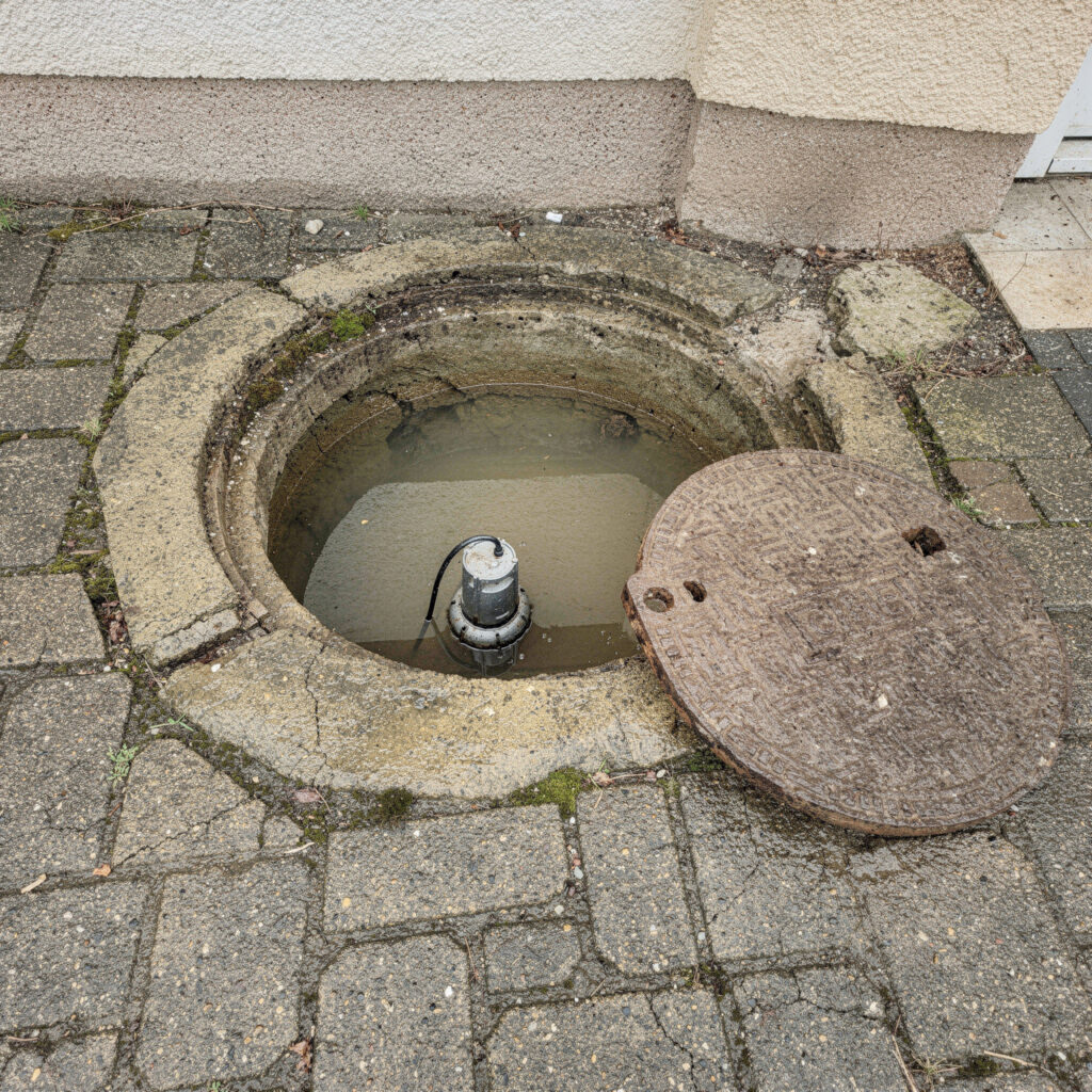 Eine unzureichende Tauchpumpe in einem geöffneten, mit trübem Wasser gefüllten Entwässerungsschacht auf Pflastersteinen.